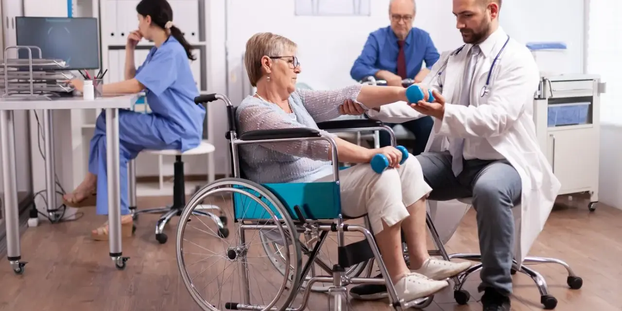 Rehabilitacja po uszkodzeniu móżdżku: pacjentka w wózku wykonuje ćwiczenia z hantlami pod okiem fizjoterapeuty.