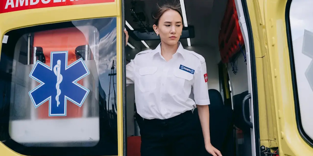 Młoda kobieta w białej koszuli z naszywką "PARAMEDICS" stoi przy otwartych drzwiach karetki. Czy kierowca karetki musi być ratownikiem?