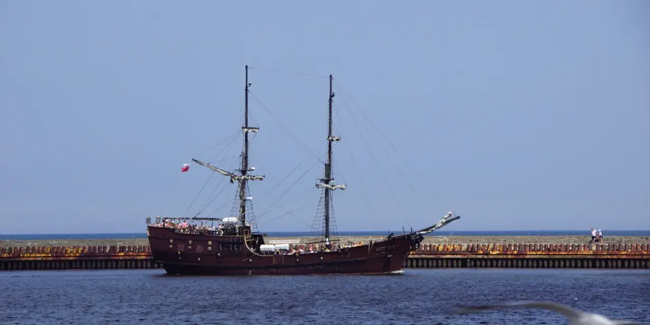 Drewniany statek z masztami i żaglami, przypominający piracki, pływa po wodzie. To tramwaj wodny Darłowo, pełen turystów.