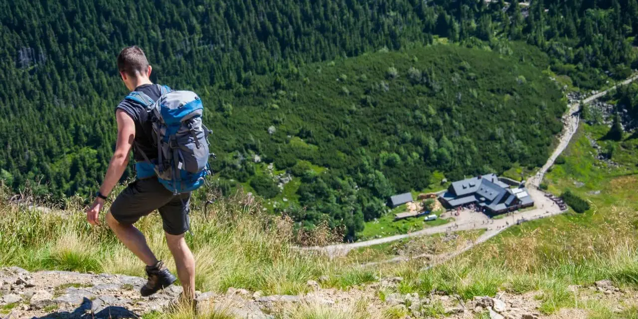 Turysta z plecakiem schodzi po kamienistej ścieżce w Karkonoszach. W dole widoczny schronisko, jedna z wielu atrakcji górskich.