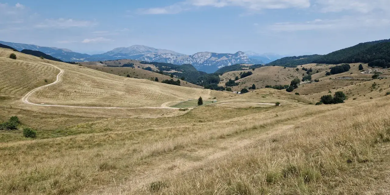Ampia vallata con erba secca e un sentiero tortuoso che sale verso il passo fittanze. Montagne sullo sfondo sotto un cielo azzurro con nuvole bianche.