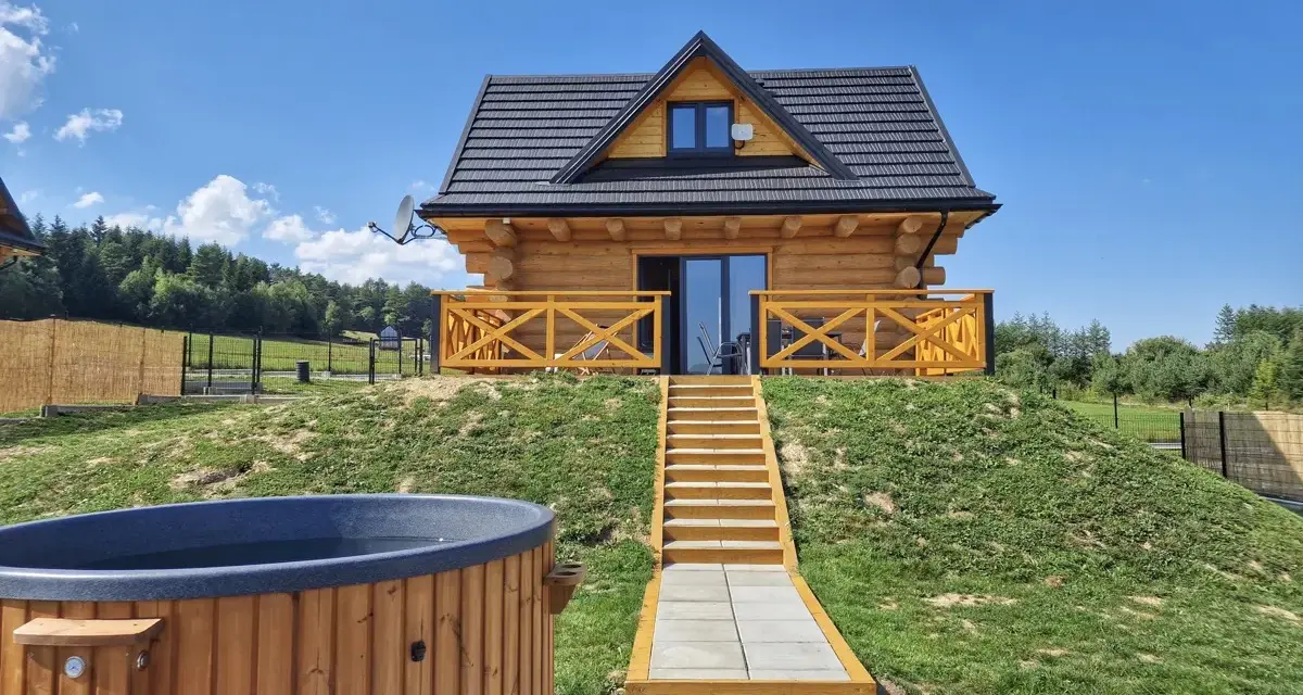 Uroczy domek na odludziu z drewnianą werandą i schodami prowadzącymi do drzwi. Przed domkiem jacuzzi i leżaki.