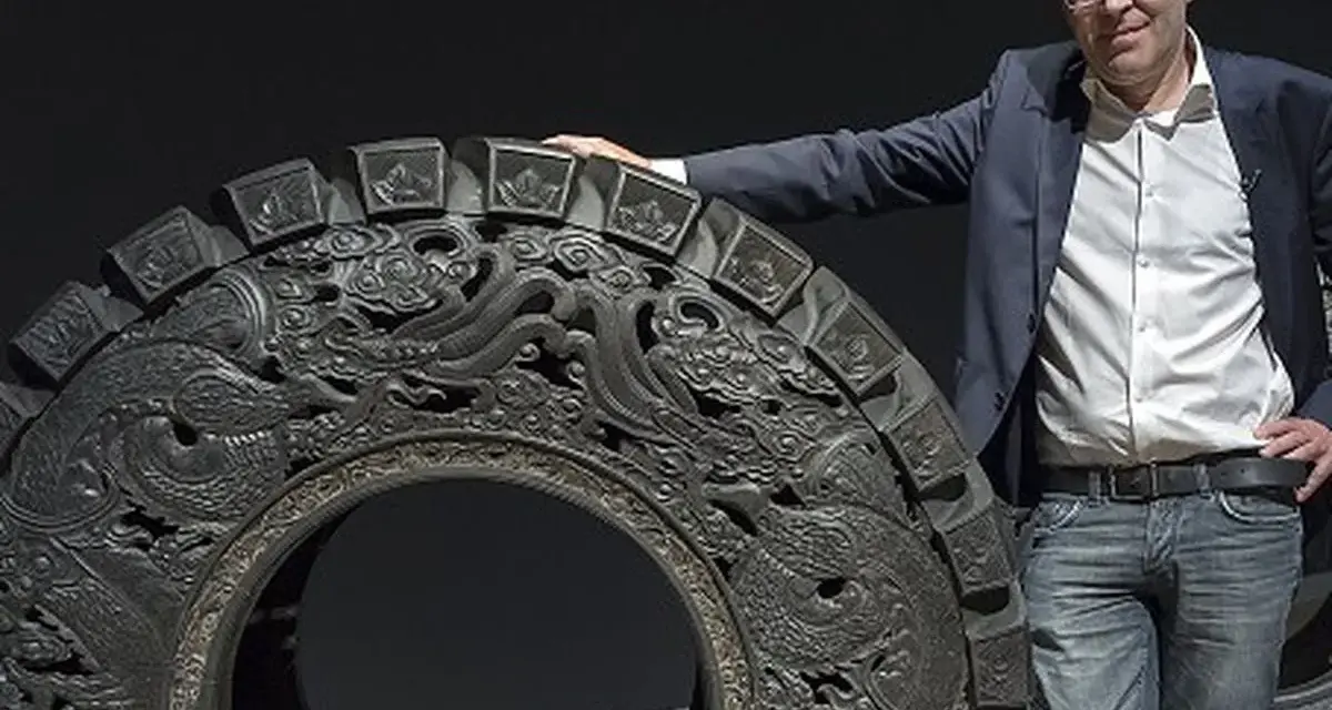 Wim Delvoye pose à côté d'une roue ornée de dragons, une œuvre d'art monumentale.