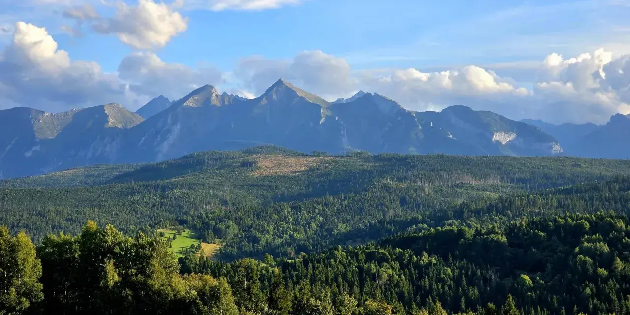 Widok na Tatry z przełęczy nad Łapszanką. Majestatyczne szczyty górskie, gęste lasy i błękitne niebo z chmurami.