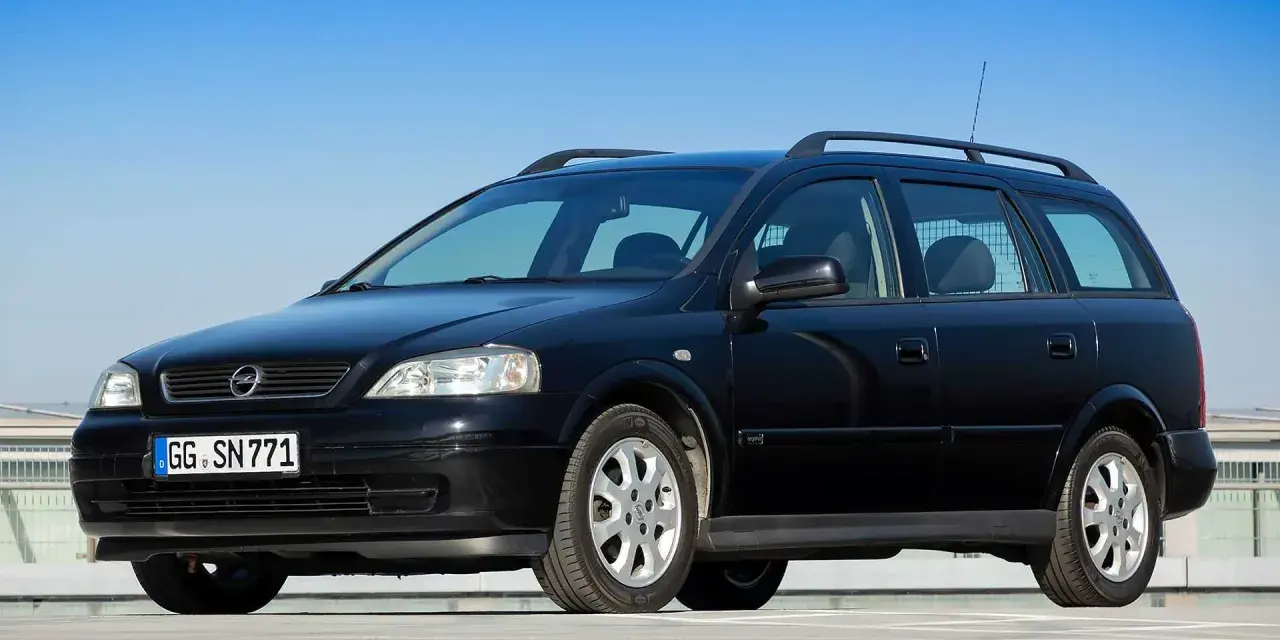 Schwarzer Opel Astra Kombi, alt, mit Dachreling und Kennzeichen GG SN 771, parkt auf einem Parkdeck vor blauem Himmel.
