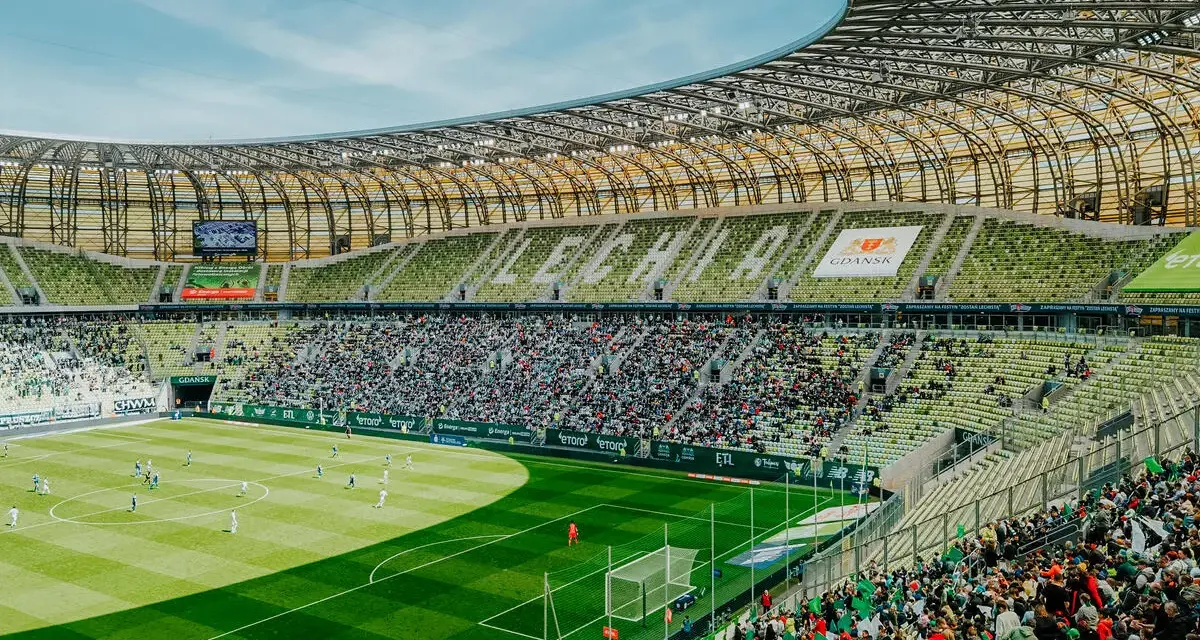 Widok na murawę i trybuny Polsat Plus Arena Gdańsk podczas meczu. Tłumy kibiców na zielonych krzesełkach.