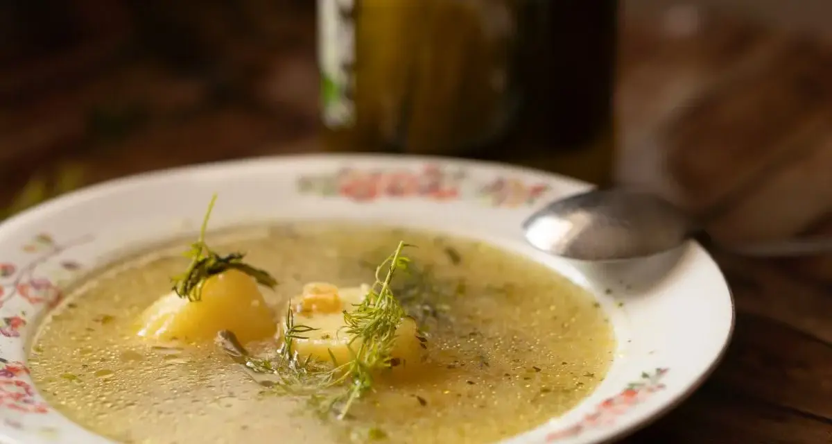 Aromatyczna keto zupa ogórkowa z ziemniakami i koperkiem, podana na talerzu z kwiatowym wzorem. Obok stoi słoik ogórków.