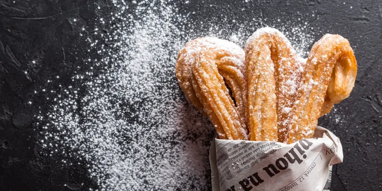 Churros z czekoladą kontra pączki z lukrem? Te złociste churros, posypane cukrem pudrem, kuszą na czarnym tle.