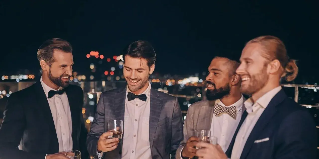 Eleganccy panowie w garniturach i muszkach, pijący drinki na tarasie. Idealny przykład, jak się ubrać na poprawiny.