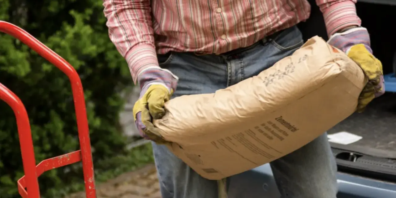 Mężczyzna w rękawicach niesie worek cementu. Zastanawia się, ile piasku na 25 kg cementu będzie potrzebne do budowy.