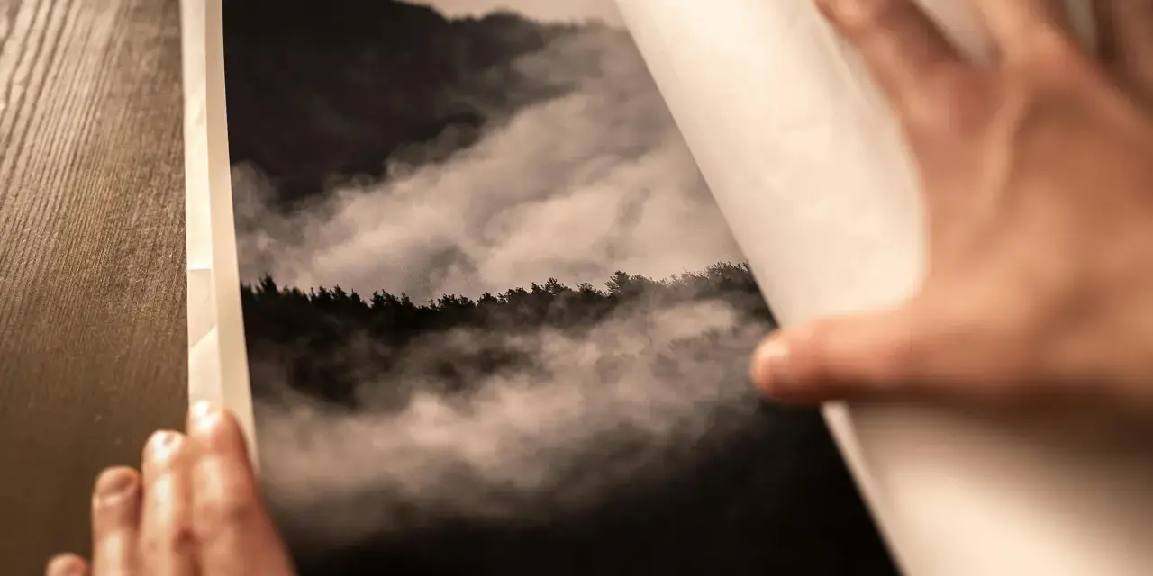 Dłonie otwierają album z fotografią górskiego lasu we mgle, wydrukowaną na papierze barytowym.