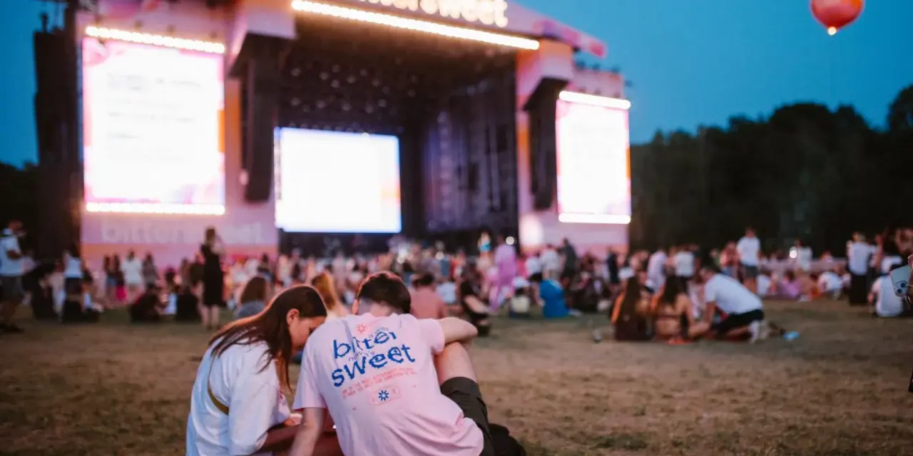 Para siedząca na trawie przed sceną festiwalową z napisem "bittersweet". W tle tłum ludzi i wielkie ekrany.