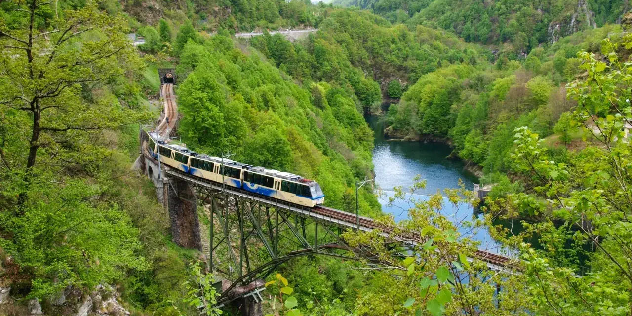 A train travels across a bridge over a river, surrounded by lush green mountains. This scenic route is part of the journey from Amsterdam to Zurich.