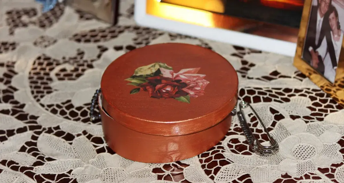 Okrągłe pudełko ozdobione techniką decoupage z motywem róż. Obok broszka i ramka ze zdjęciem.