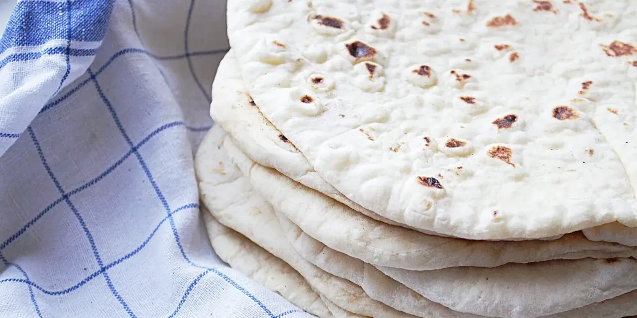 En hög med nybakade tortillabröd, perfekta för ditt nästa tortilla recept. Bredvid ligger en blåvitrutig kökshandduk.
