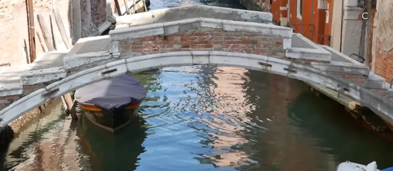 Un piccolo ponte a Venezia, senza parapetto, sovrasta un canale dove una barca coperta attende.