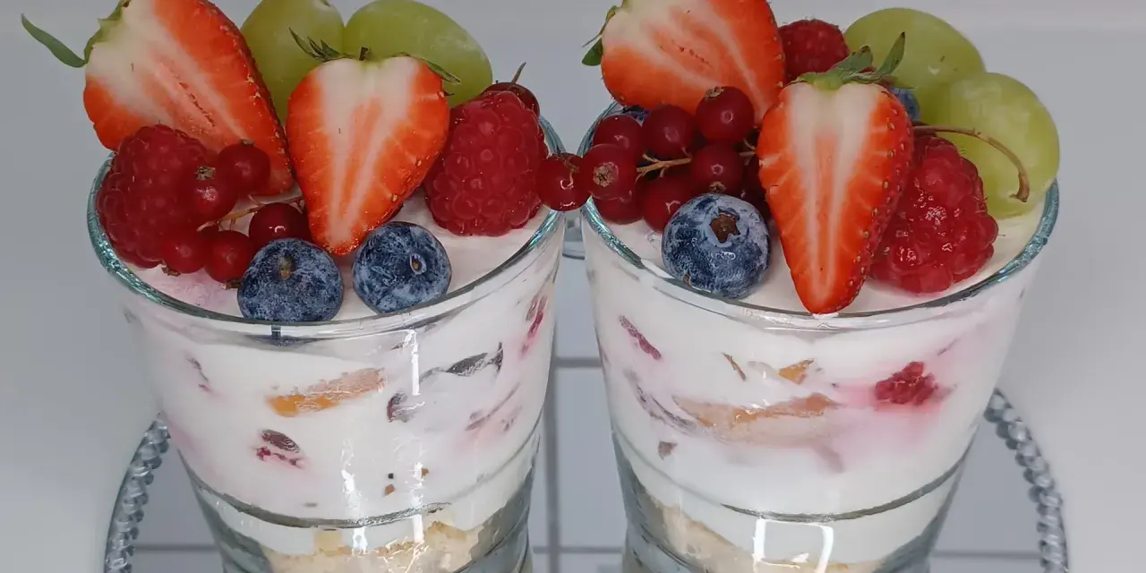 Zwei Gläser mit einem cremigen Dessert, garniert mit frischen Erdbeeren, Himbeeren, Blaubeeren, Johannisbeeren und Trauben. Ein köstliches Dessert im Glas.