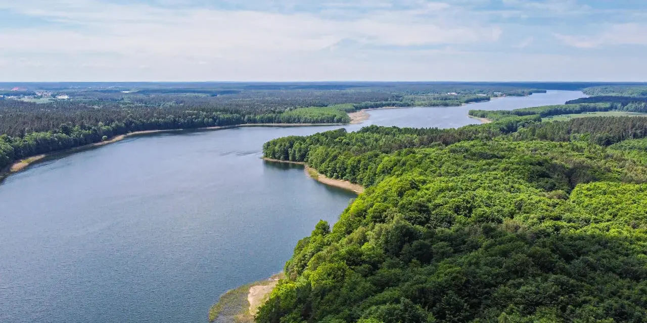 Malownicze jezioro otoczone lasem, idealne miejsce na nietypowe atrakcje Mazury. Widok z lotu ptaka na spokojną taflę wody i bujną zieleń.
