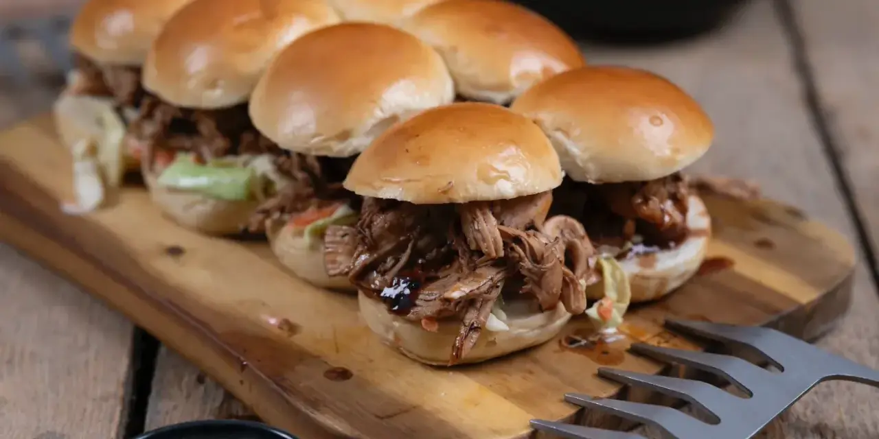 Saftiges pulled chicken im Backofen zubereitet, serviert in kleinen Brötchen auf einem Holzbrett.