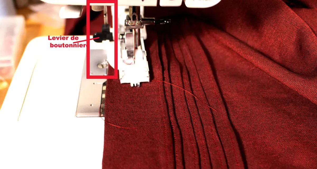 Levier de boutonnière sur machine à coudre, prêt pour réaliser une boutonnière parfaite sur un tissu bordeaux plissé.