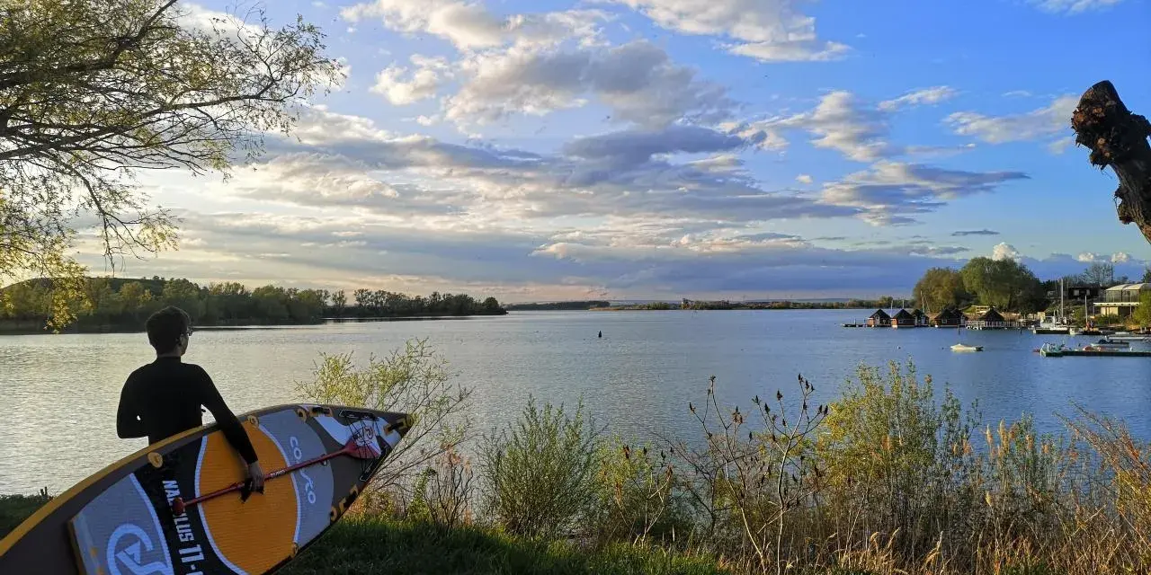 Ein junger Mann mit SUP-Board am Ufer des Alperstedter Sees. Der Himmel ist blau mit weißen Wolken, die Sonne scheint.