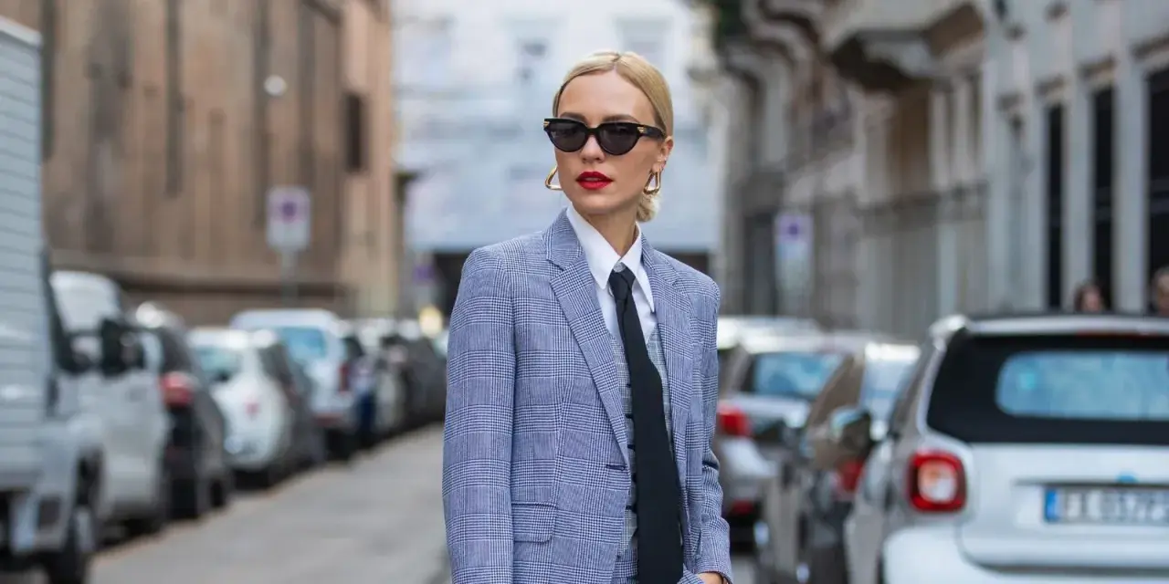 Mujer con traje a cuadros, camisa blanca y corbata negra, luciendo un impecable tomboy style. Gafas de sol y labios rojos completan su look.
