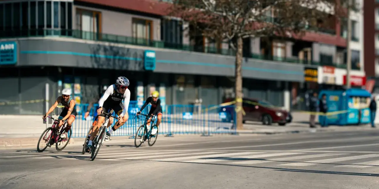 Cyclists race through a city street, perhaps part of le grand tour de France, with buildings and spectators lining the course.