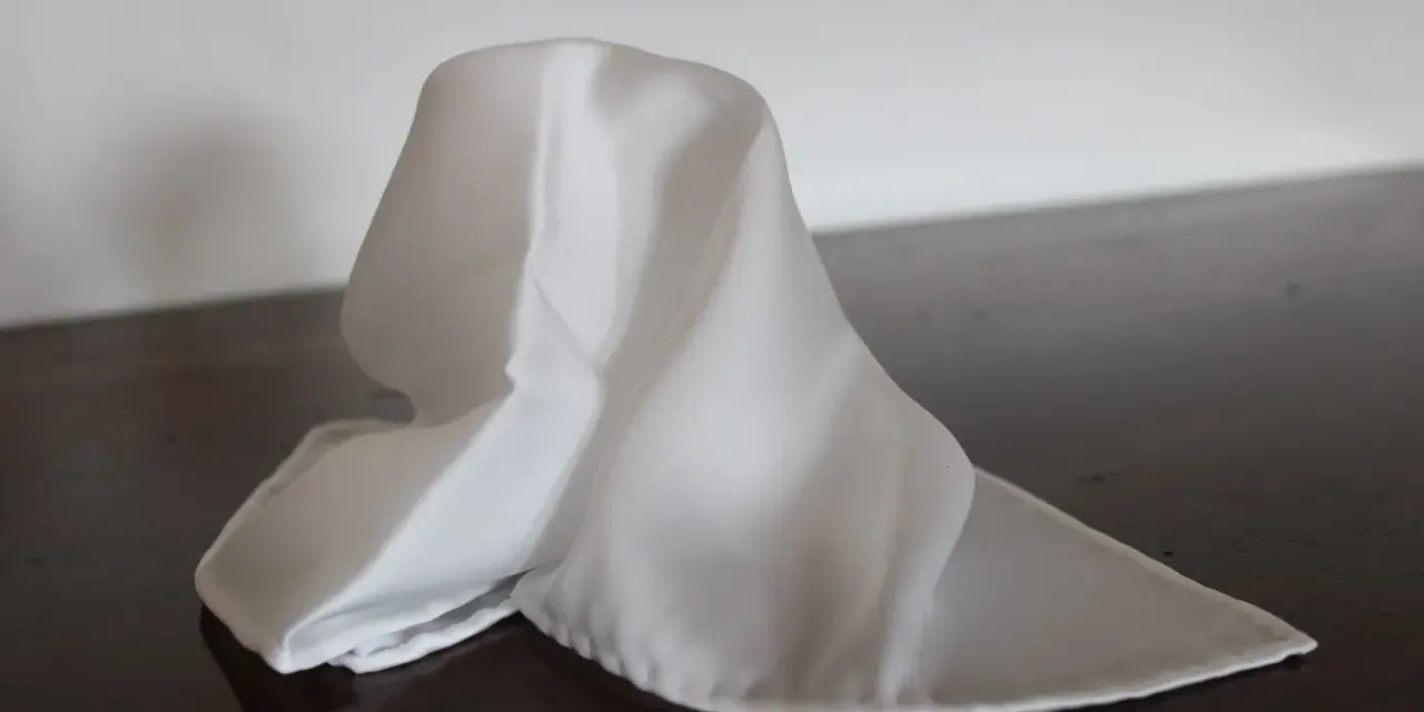 Un fazzoletto bianco di seta, drappeggiato su un tavolo scuro. Il suo significato è legato alla purezza e all'eleganza.