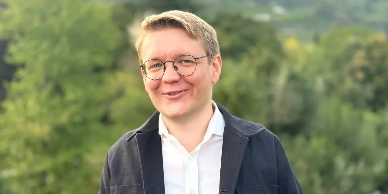 Ein Mann mit Brille lächelt in die Kamera. Er trägt ein weißes Hemd und eine dunkle Jacke. Im Hintergrund sind Bäume und Berge zu sehen. Er studiert Seelsorge.