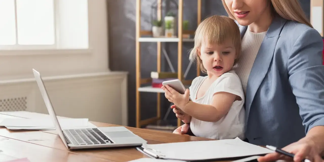 Pracująca mama z dzieckiem na kolanach, laptop i tablet na biurku. Czy na macierzyńskim można pracować? Tak, to możliwe!