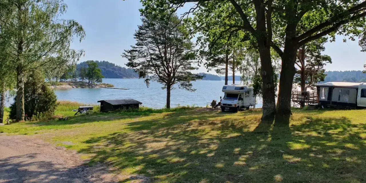 Solig dag vid vattnet på Kalvholmens camping. Husbilar och husvagnar står uppställda på gräsmattan nära sjön.