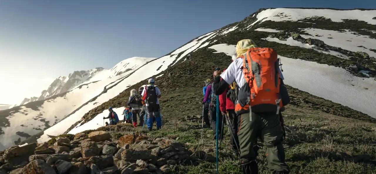 Un groupe de randonneurs progresse sur un sentier rocailleux, le soleil éclairant les montagnes enneigées. Ils semblent déterminés à savoir son altitude.