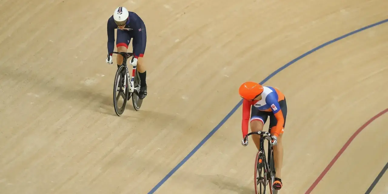 Two cyclists compete in a track sprint. One rider in a blue and white uniform leads, while another in orange and blue follows closely.