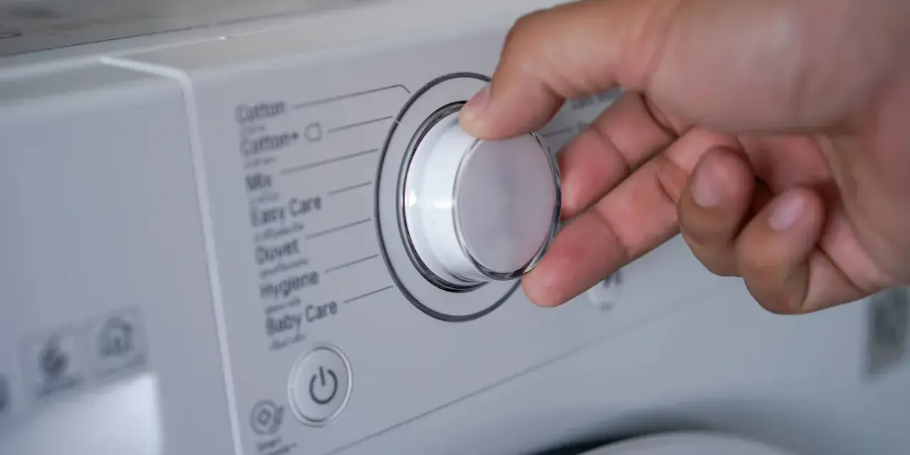 Mano ajustando el selector de la lavadora para saber como lavar un anorak. Se ven programas como "Cotton", "Mix", "Duvet".