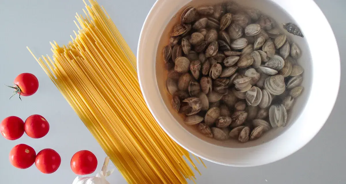 Składniki na pyszny makaron z małżami: spaghetti, pomidorki, czosnek i świeże małże w misce.