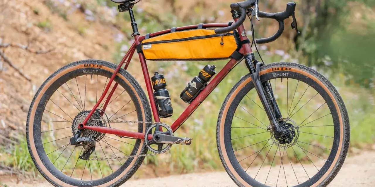 A red gravel bike with an orange frame bag and two water bottles. This bike, like many gravel bikes, does not have suspension, relying on its tires and frame for comfort.