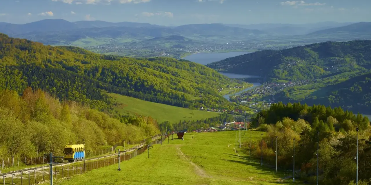 Widok na zielone **beskid mały szczyty** z kolejką linową i jeziorem w oddali.