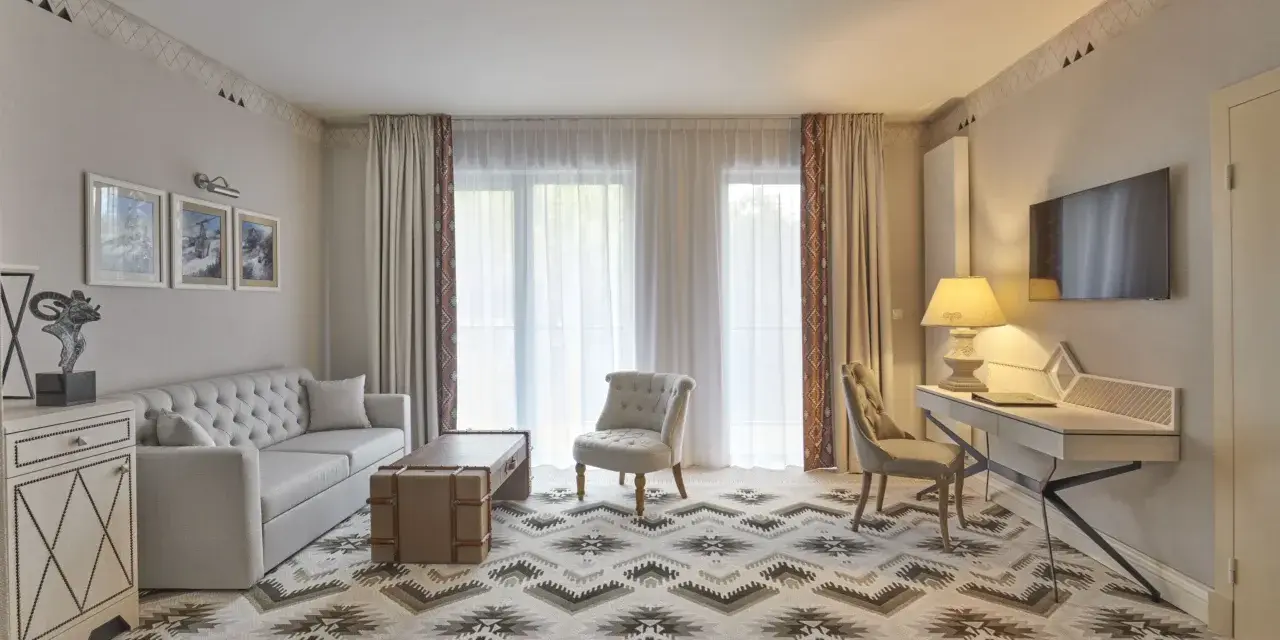 Elegancki pokój w hotelu 5 gwiazdkowym: sofa, stolik, fotele, biurko i TV. Wzory na dywanie i zasłonach dodają charakteru.