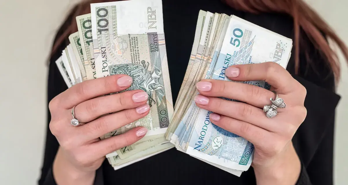 Dłonie z paznokciami w kolorze nude trzymają stosy banknotów 100 i 50 zł. To idealna jubileuszówka na prezent!