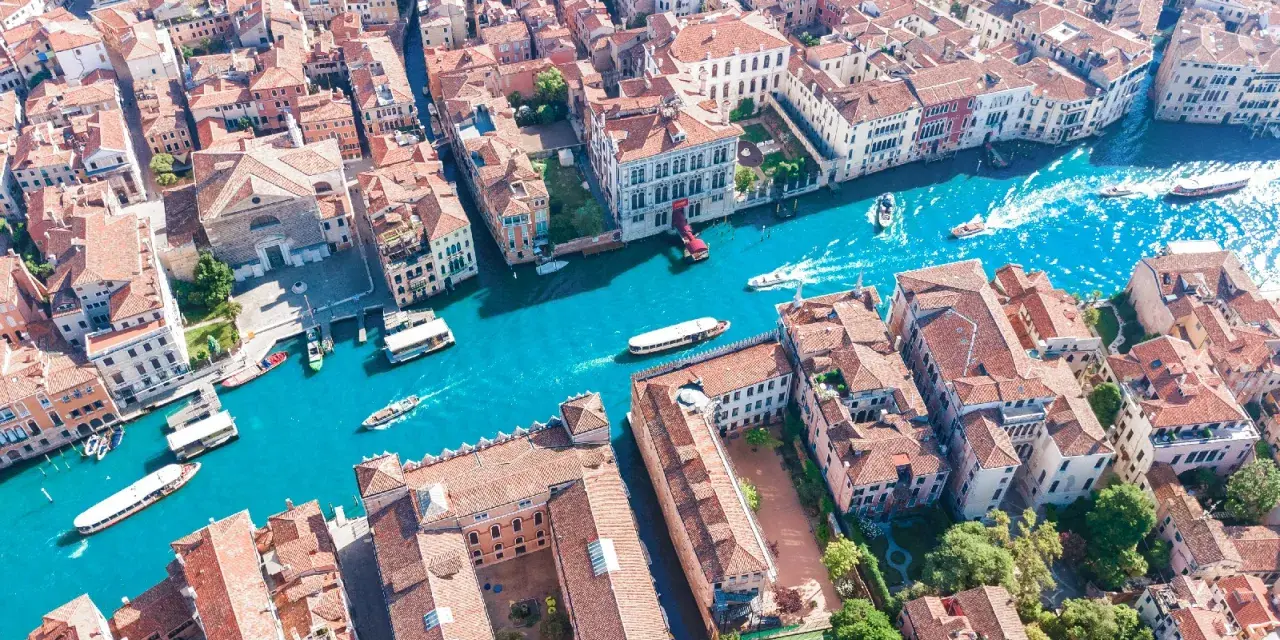 Vista aerea del **canale di Venezia** con gondole e vaporetti che navigano tra palazzi storici dai tetti rossi.