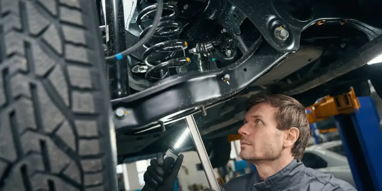 Mechanik bada elementy zawieszenia samochodu, oświetlając je latarką. Widoczna sprężyna i amortyzator.