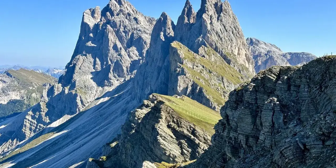 Majestätische Felszinnen ragen in den blauen Himmel. Eine **Seceda Wanderung** führt durch grüne Almwiesen und felsiges Gelände.