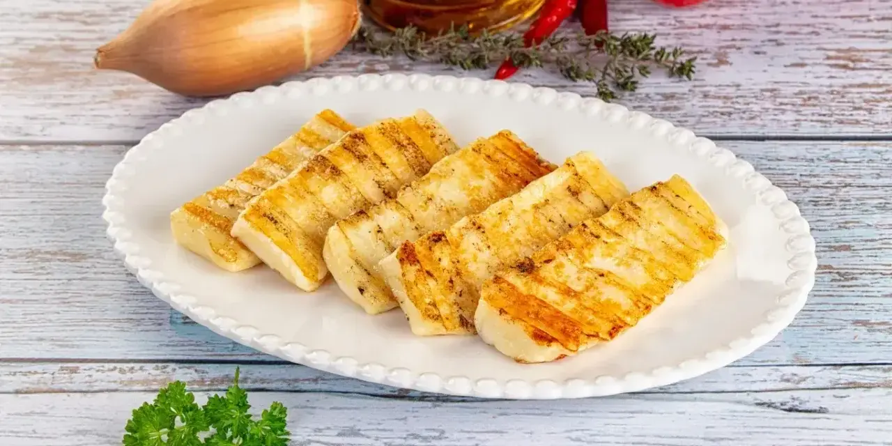Grillowany ser halloumi na białym talerzu, z dodatkami jak czosnek, cebula i pomidory w tle.