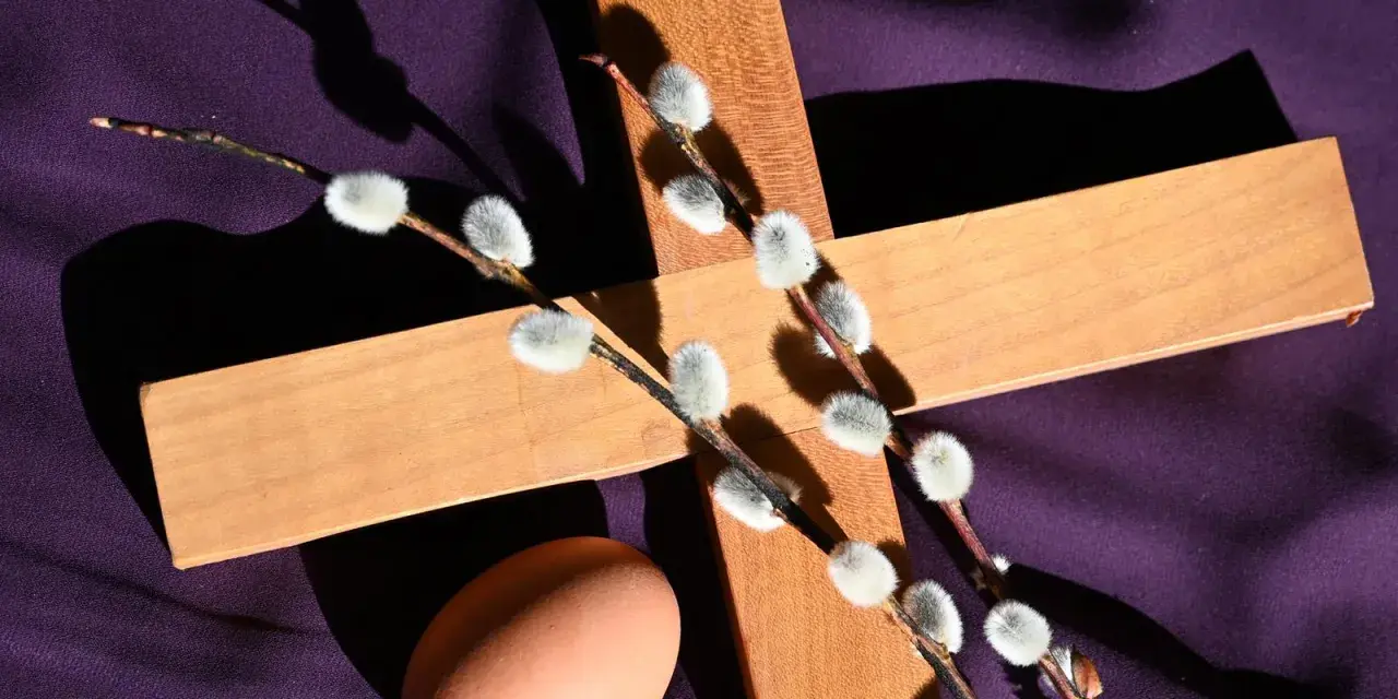 Holzkreuz mit Weidenkätzchen und Ei auf lila Stoff, ein Symbol für christliche Ostern.