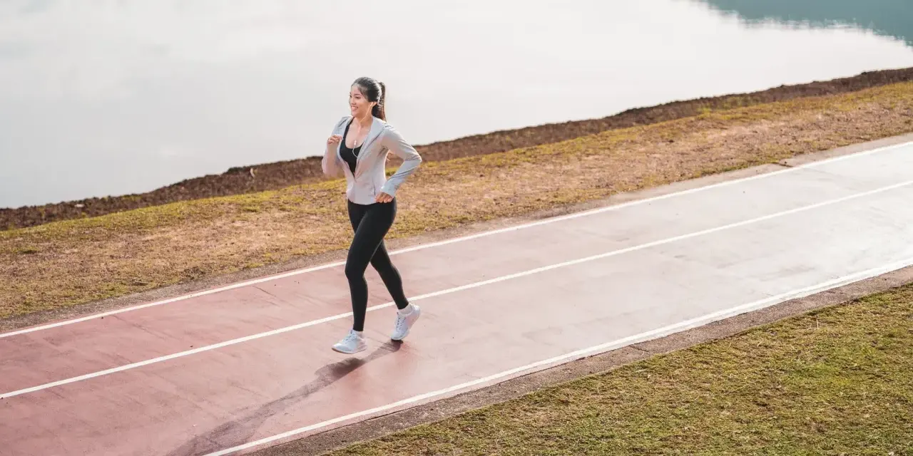 Une femme souriante profite d'une marche rapide sur une piste de course, au bord d'un lac paisible.