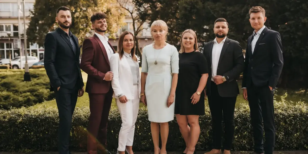 Grupa sześciu profesjonalistów w eleganckich strojach stoi przed budynkiem i zielenią.