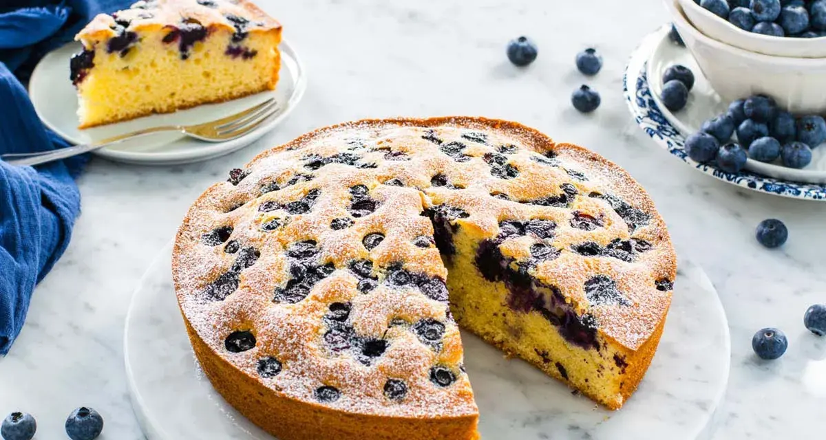 Deliziosa torta allo yogurt con mirtilli, spolverata di zucchero a velo. Una fetta è già servita su un piattino.