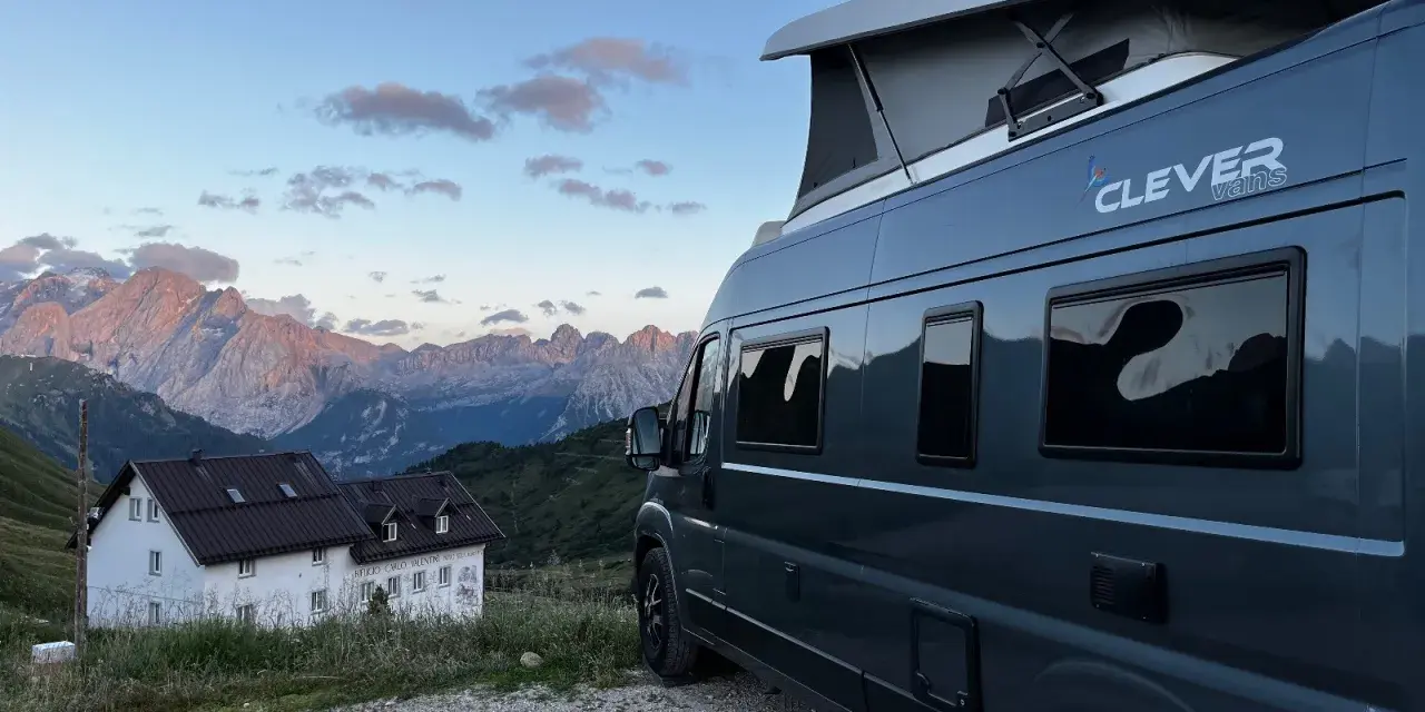 En mörkgrå husbil med pop-up tak, märkt "Clever vans", står parkerad vid en bergig stuga med Alperna i bakgrunden.