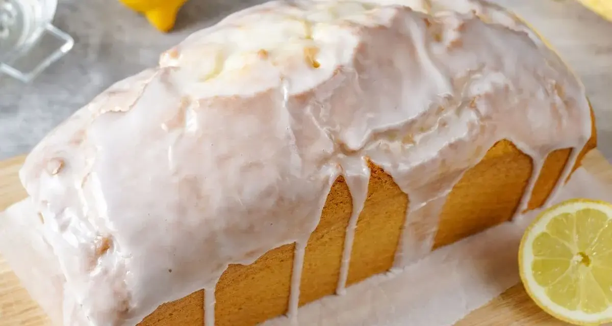 Saftiger zitronenkuchen in Kastenform, überzogen mit süßem Zuckerguss. Ein Zitronenstück liegt daneben.