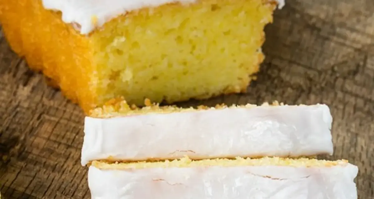 Fetta di plumcake al limone con glassa bianca e una fetta di limone sopra, su un tagliere di legno.